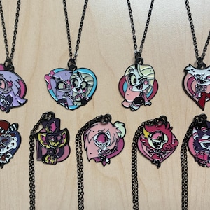 May include: Collection of ten necklaces with black chains and character pendants. The pendants feature colorful cartoon characters in various shapes, including hearts and stylized figures. The color palette includes pink, purple, and blue.