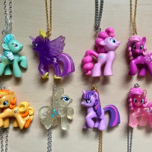 Puede incluir: Ocho collares con colgantes de ponis de colores. Los ponis son de diferentes colores, incluyendo verde, morado, rosa, naranja y transparente. Los collares tienen cadenas plateadas y doradas.