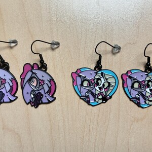 May include: Four pairs of dangle earrings. Two pairs feature a cartoon character's head in shades of purple, pink, and black. The other two pairs are heart-shaped, with a cartoon character in shades of purple, pink, and blue.