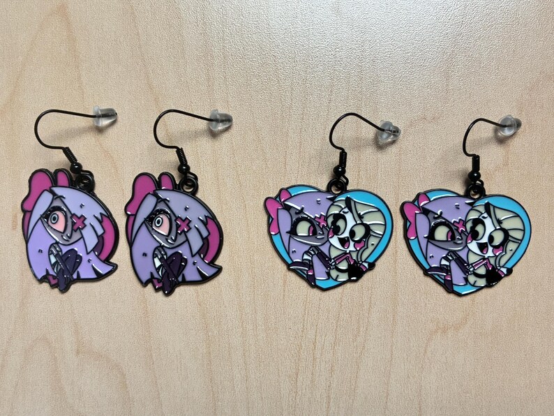 May include: Four pairs of dangle earrings. Two pairs feature a cartoon character's head in shades of purple, pink, and black. The other two pairs are heart-shaped, with a cartoon character in shades of purple, pink, and blue.