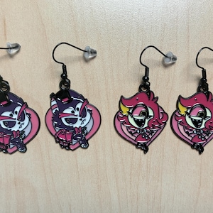 May include: Four pairs of dangle earrings with black hooks and clear stoppers. Two pairs feature a purple cat design with pink accents, and two pairs showcase a pink character with yellow details. All earrings have a black outline.