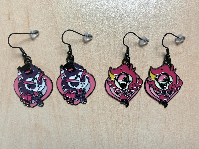 May include: Four pairs of dangle earrings with black hooks and clear stoppers. Two pairs feature a purple cat design with pink accents, and two pairs showcase a pink character with yellow details. All earrings have a black outline.