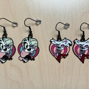 May include: Four pairs of black dangle earrings. Two pairs feature a cartoon character with a pink, blue, and white color scheme. The other two pairs are heart-shaped with a white character and red accents.