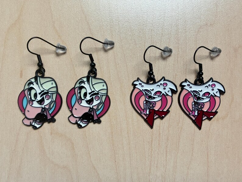 May include: Four pairs of black dangle earrings. Two pairs feature a cartoon character with a pink, blue, and white color scheme. The other two pairs are heart-shaped with a white character and red accents.