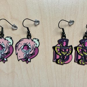 May include: Four pairs of dangle earrings. Two pairs feature pink cartoon characters with white eyes and hair, and the other two pairs feature purple and yellow cartoon characters. All earrings have black hooks and clear earring backs.