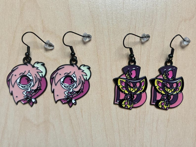 May include: Four pairs of dangle earrings. Two pairs feature pink cartoon characters with white eyes and hair, and the other two pairs feature purple and yellow cartoon characters. All earrings have black hooks and clear earring backs.