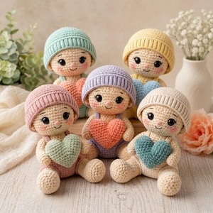 May include: Five handmade crochet dolls, each holding a heart. The dolls wear pastel-coloured hats and dungarees. The hearts are in shades of pink, green, and blue. The dolls are sitting on a light-coloured wooden surface.