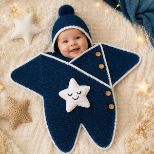 May include: A dark blue crocheted star-shaped baby bunting with a matching hat featuring a pom-pom. The bunting has white trim, three brown wooden buttons along the side, and a white crocheted star appliqué with a stitched smiling face.