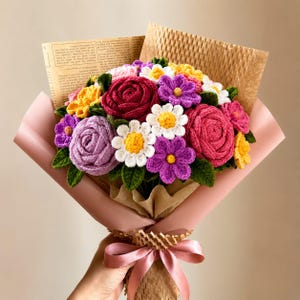 Op de afbeelding: Een handgemaakt boeket van gehaakte bloemen in verschillende kleuren, waaronder rood, roze, paars, geel en wit. De bloemen zijn verpakt in bruin papier en roze tissuepapier, vastgebonden met een roze lint. Het boeket bevat rozen, madeliefjes en andere bloemmotieven.