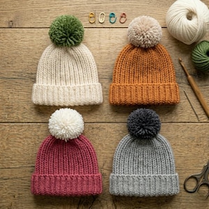 May include: Four knitted beanies in cream, orange, pink, and gray, each topped with a matching or contrasting pom-pom. The hats are arranged on a wooden surface, with knitting supplies visible.