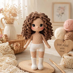May include: A handcrafted crochet doll with brown, curly hair, rosy cheeks, and large black eyes. The doll is dressed in white shorts and white boots. The background features yarn, a wooden heart with text, and a vase of flowers.