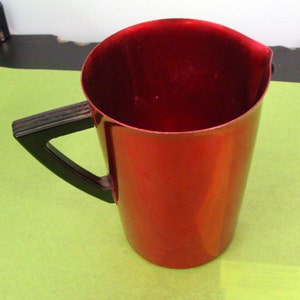 May include: A red metal pitcher with a black plastic handle. The pitcher is empty and has a smooth, shiny surface.