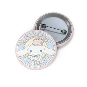 May include: Two round, pink Cinnamoroll badges. One badge features a white cartoon dog with blue eyes, a teacup hat, and a bow. The word "Cinnamoroll" is written around the dog. The other badge shows the pin back.