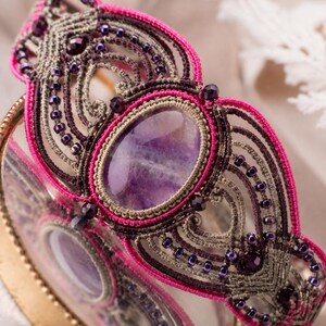Può includere: Un bracciale in macramè con una grande pietra ovale di ametista viola al centro, circondata da intricati nodi nelle tonalità del rosa acceso, marrone scuro e beige. Il design è arricchito da piccole perline viola scuro e dal testo Amara Roho.