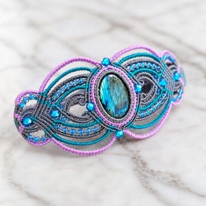 May include: A macrame hair clip featuring a central oval labradorite stone with blue and gold iridescence. The accessory is crafted with grey, teal, and lavender cord, accented with small blue faceted beads arranged in a symmetrical pattern.