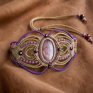 Könnte beinhalten: Ein Makramee-Armband mit einem zentralen, ovalen, durchscheinenden violetten Stein, eingefasst in beigefarbene und violette Kordeln. Das Design umfasst komplexe Knotenmuster, kleine violette Perlen und verstellbare Bänder mit dunklen Perlen.