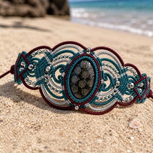 Op de afbeelding: Een macramé armband met een centrale ovale donkergrijze steen met een stippenpatroon. De armband is gemaakt van bordeauxrood, groenblauw en lichtgrijs koord, geaccentueerd met kleine metallic zilveren kralen in het vlechtwerk.