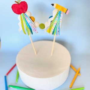 May include: Two cake toppers, one with a red felt apple and the other with a yellow felt pencil, both with colorful ribbons and alphabet letters. The toppers are on a white cake stand with colorful pens scattered around.