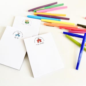 May include: Three white notepads with different designs. The top notepad has a green and yellow house with the text "From the classroom of Mrs. [unclear]" The middle notepad has a pair of blue scissors with the text "From the office of [unclear] & [unclear]" The bottom notepad has a red schoolhouse with the text "From the classroom of Mrs. Carol". There are multiple colored markers scattered around the notepads.