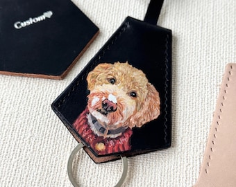 May include: Black leather keychain featuring a hand-painted portrait of a dog with tan fur and a red sweater. The keychain has a silver key ring and a black leather strap. The word "Custom" is printed on a separate black leather piece.