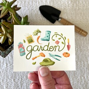 May include: A white card with the word "garden" written in green cursive. The card is surrounded by illustrations of gardening tools and plants, including a pair of gloves, a watering can, a trowel, a pair of pruning shears, a hat, a tomato, a carrot, a seed packet, a boot, and a vine.
