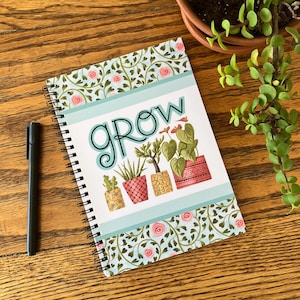 May include: A spiral-bound notebook with the word "GROW" and illustrations of potted plants. The cover features a floral design with pink roses and green leaves. A black pen rests beside the notebook on a wooden surface.