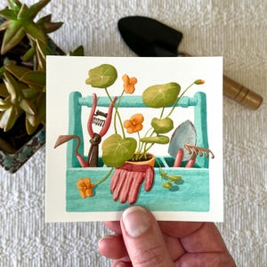 May include: A blue gardening tool caddy with a variety of tools, including a trowel, pruning shears, and a hand rake. A potted plant with orange flowers is in the caddy.