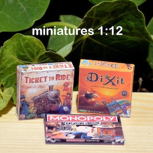 Op de afbeelding: Drie miniatuur bordspellen: Ticket to Ride, Dixit en Monopoly. De Ticket to Ride doos is bruin met een trein op de voorkant. De Dixit doos is oranje met een fantasierijke illustratie. De Monopoly doos is rood met een cartoonfiguur op de voorkant.