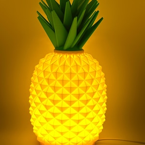 Könnte beinhalten: Gelbe Ananas-förmige Lampe mit geometrischem Muster und grünen Blättern. Die Lampe leuchtet und strahlt ein warmes Licht aus. Die Lampe ist mit einem weißen Kabel angeschlossen. Die Lampe ist eine dekorative Leuchte.