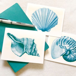May include: Three watercolor paintings of seashells on white card stock. The shells are painted in shades of blue and green. The shells are a scallop, a conch, and a nautilus.