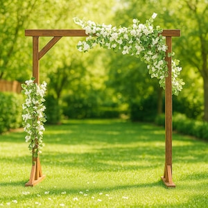 Puede incluir: Un arco de boda de madera adornado con flores blancas. El arco de madera marrón está decorado con flores blancas y follaje. Se encuentra sobre un césped verde, con árboles en el fondo, creando un entorno natural.