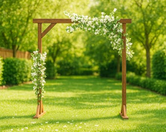 May include: A wooden wedding arch with white floral accents. The arch is constructed from brown wood and is decorated with white flowers and greenery. The arch is set up on a green lawn, with trees in the background.