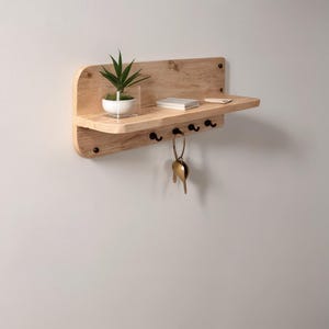 May include: A light-colored wooden wall shelf with a small shelf, four hooks, and a small plant in a white bowl. The shelf holds a small stack of white notepads and a gold-colored set of keys.