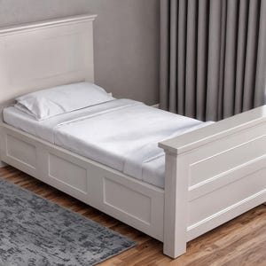 May include: A light-colored, wooden twin bed with a headboard and footboard. The bed is made up with white sheets and a pillow. A gray rug is in front of the bed, and the floor is wood.