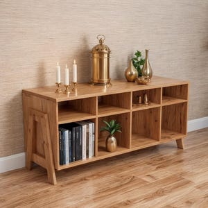 May include: A light oak wood cabinet with multiple cubby shelves. The top displays gold decor, including a lantern, vases, and candles. Books and a small plant are visible in the shelves. The cabinet is on a wood floor.