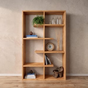 May include: A light brown wooden bookcase with various shelves and compartments. The shelves display decorative items such as books, plants, bottles, and a decorative clock. The bookcase is set against a neutral-toned wall.