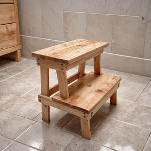 May include: A two-tier wooden step stool with a natural finish. The stool has a rectangular top and a lower step, with visible screw fixings. The wood grain is visible. The stool is designed for use in a bathroom or similar setting. The dimensions are approximately 40cm x 30cm x 30cm.