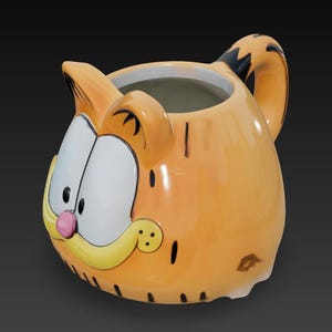 Puede incluir: Taza de cerámica naranja con forma de cabeza de Garfield. La taza presenta la cara de Garfield con ojos blancos, nariz rosa y bigote amarillo. El asa está diseñada para parecerse a la cola rayada de Garfield. Un artículo divertido y novedoso.