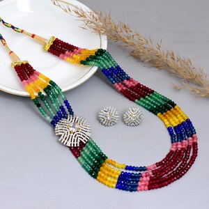 May include: A multi-strand necklace and matching earrings set. The necklace features strands of colorful beads in red, pink, yellow, green, and blue. A silver-toned, flower-shaped pendant with clear stones is the focal point. The earrings are small, round, and also feature clear stones.