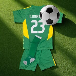 Op de afbeelding: Een groen en geel voetbaltenue met het nummer 23 en de naam "E. MART" op de rug. Een voetbal met zwart-witte panelen is boven het tenue geplaatst. Een groene sok met grijze accenten is ook inbegrepen.