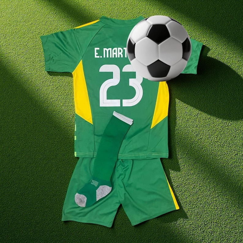 Op de afbeelding: Een groen en geel voetbaltenue met het nummer 23 en de naam "E. MART" op de rug. Een voetbal met zwart-witte panelen is boven het tenue geplaatst. Een groene sok met grijze accenten is ook inbegrepen.