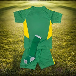 May include: A green and yellow soccer uniform set, including a short-sleeved jersey, shorts, and a single sock. The jersey and shorts are green with yellow accents. The sock is green with gray details. The set is displayed on a grassy field.