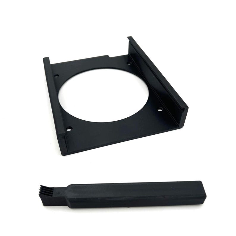 Puede incluir: Soporte met&aacute;lico negro con una abertura circular y orificios de montaje, y un componente rectangular negro separado con un extremo en forma de cepillo. Los art&iacute;culos se muestran sobre un fondo blanco.