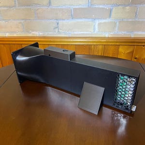 Peut inclure: Un appareil rectangulaire noir avec un ventilateur et un petit panneau noir s&eacute;par&eacute;. L'appareil a une forme carr&eacute;e avec une entr&eacute;e incurv&eacute;e et un &eacute;vent en nid d'abeille. L'appareil est pos&eacute; sur une surface en bois.