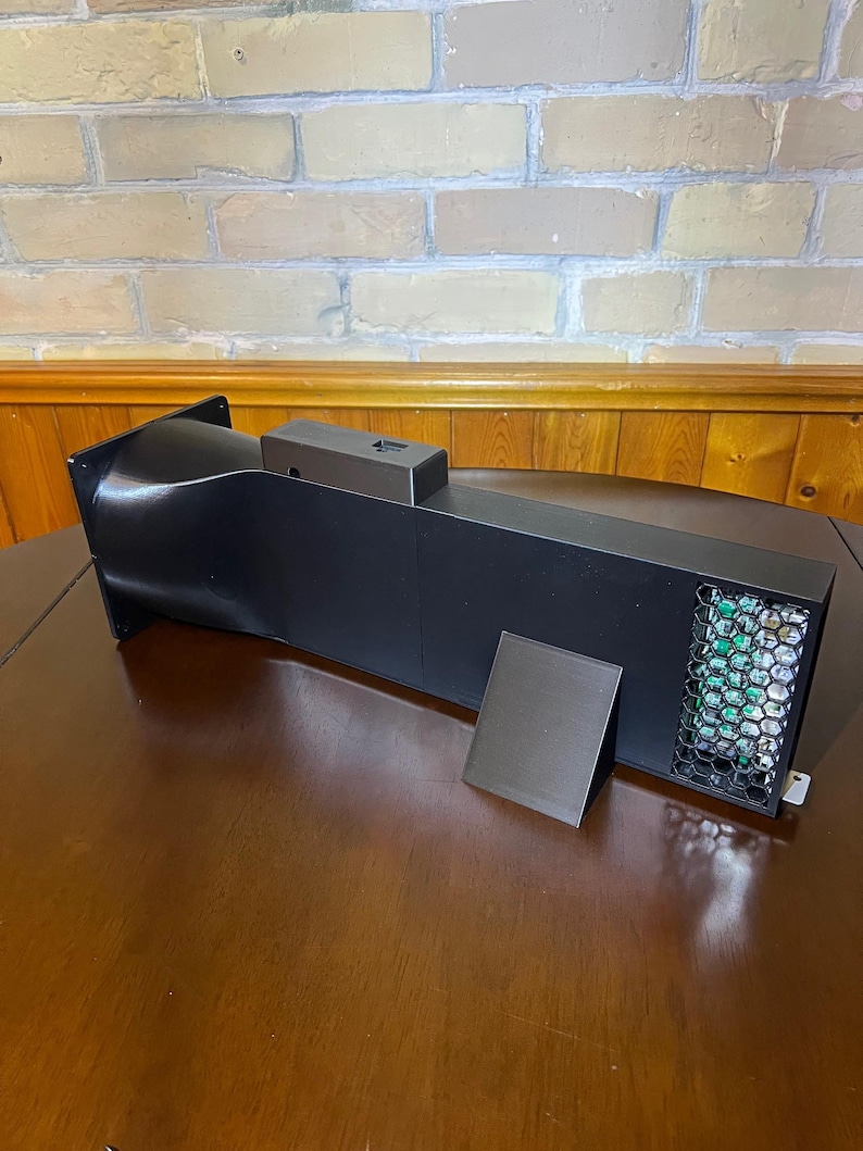 Peut inclure: Un appareil rectangulaire noir avec un ventilateur et un petit panneau noir s&eacute;par&eacute;. L'appareil a une forme carr&eacute;e avec une entr&eacute;e incurv&eacute;e et un &eacute;vent en nid d'abeille. L'appareil est pos&eacute; sur une surface en bois.