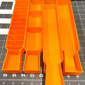 Puede incluir: Organizador de plástico naranja brillante con múltiples compartimentos y bandejas. El organizador tiene secciones rectangulares y cuadradas de varios tamaños, ideales para guardar objetos pequeños. El diseño incluye varias bandejas largas y estrechas y secciones más pequeñas.