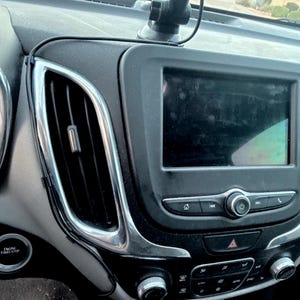 May include: A black car dashboard centre console featuring a rectangular infotainment touchscreen, a vertical air vent with chrome trim, a power button, and climate control knobs. A black cable runs along the side of the console unit.