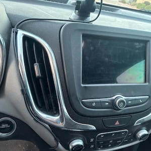 Puede incluir: Una consola central de tablero de automóvil negra que presenta una pantalla táctil de infoentretenimiento rectangular, una rejilla de ventilación vertical con moldura cromada, un botón de encendido, botones multimedia y perillas de control de clima.