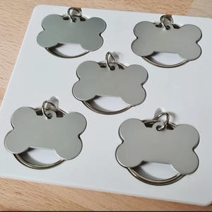 May include: Five silver bone-shaped pet ID tags with metal rings, displayed on a white tray. The tags are blank, ready for engraving. The tray is on a wooden surface. Each tag is approximately 4 cm long.