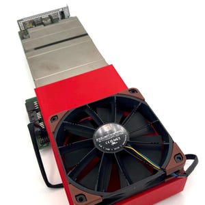 Pode incluir: Um componente de computador com uma ventoinha preta e detalhes castanhos, montado numa base vermelha. Inclui um dissipador de calor prateado e uma placa de circuito verde. A ventoinha tem o texto "industriapPC".